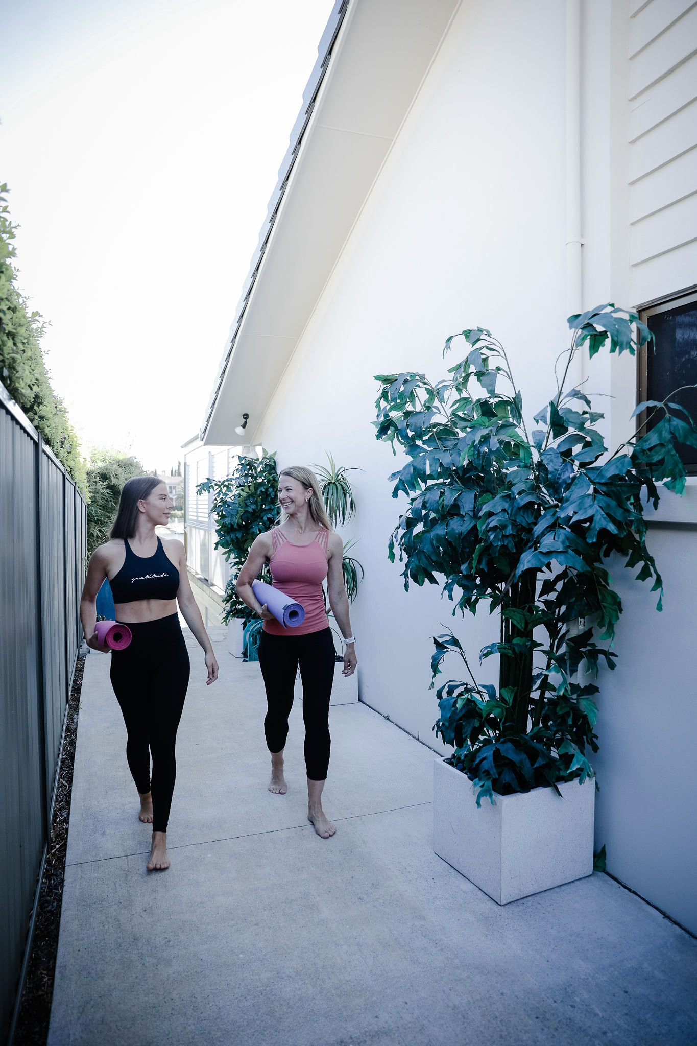 Two women walking barefoot along a sunlit path beside a house, rolled exercise mats under their arms, talking as they go
