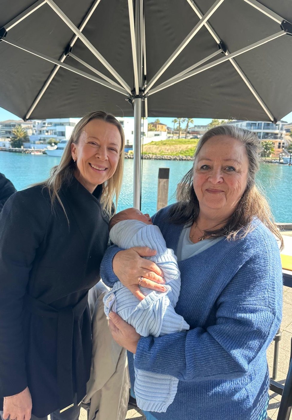 Miranda holding her great-grandson, with her daughter Rouxchelle beside her at a waterfront cafe